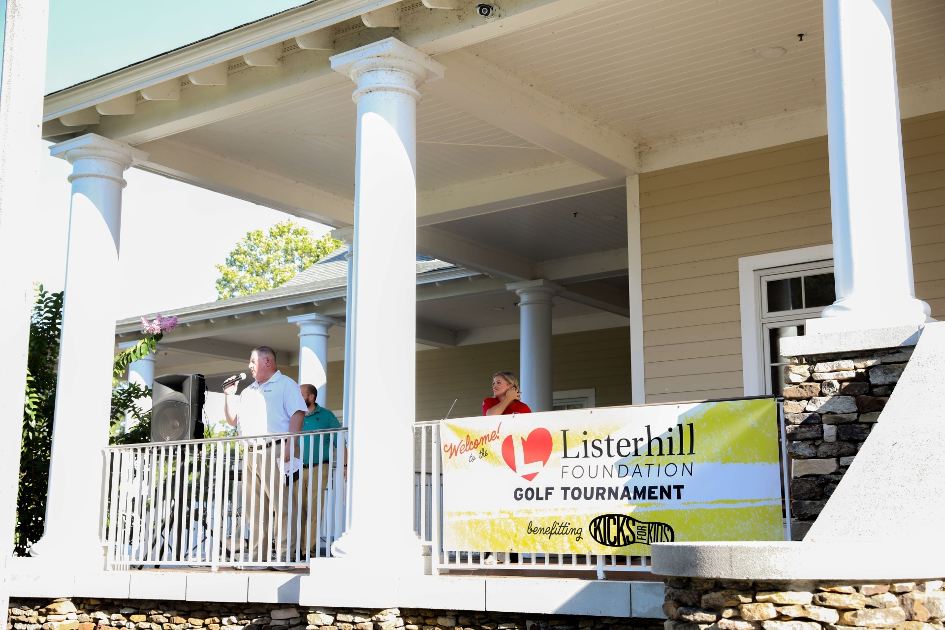 Brad Green speaks into a microphone while standing on the porch of RTJ's club house addressing a crowd of golfers. Haley Brink, Director of Philanthropy, stands behind him and sign saying "Welcome to the Listerhill Foundation Golf Tournament benefitting Kicks for Kids".